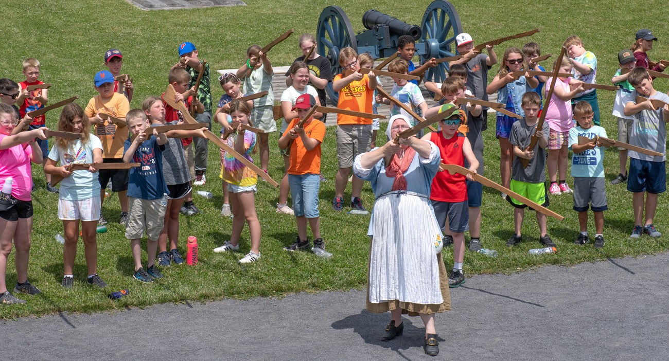 Dozens of children in lines behind a woman in 18th C clothing. They are all holding and pointing wooden muskets in the air.