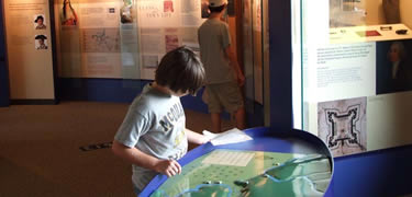 A young man stares at a blue and green map, the sun reflects off of the shiny textured surface as he traces it