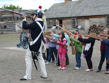a soldier in a tall hat directs young girls how to stand in a postion of a soldier while holding a musket. all are in a neat row