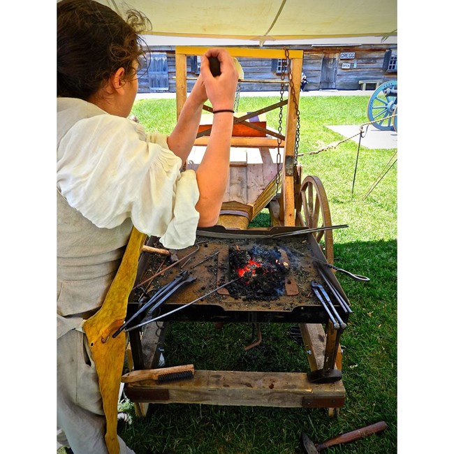 Looking over the shoulder of a person pulling a wooden handle connected to a forge bellows. There is a tray filled with fiery embers in front of them.