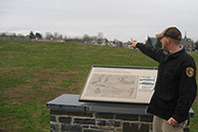 near a sign, a man points towards the fort over a green hill on a coudly grey day.