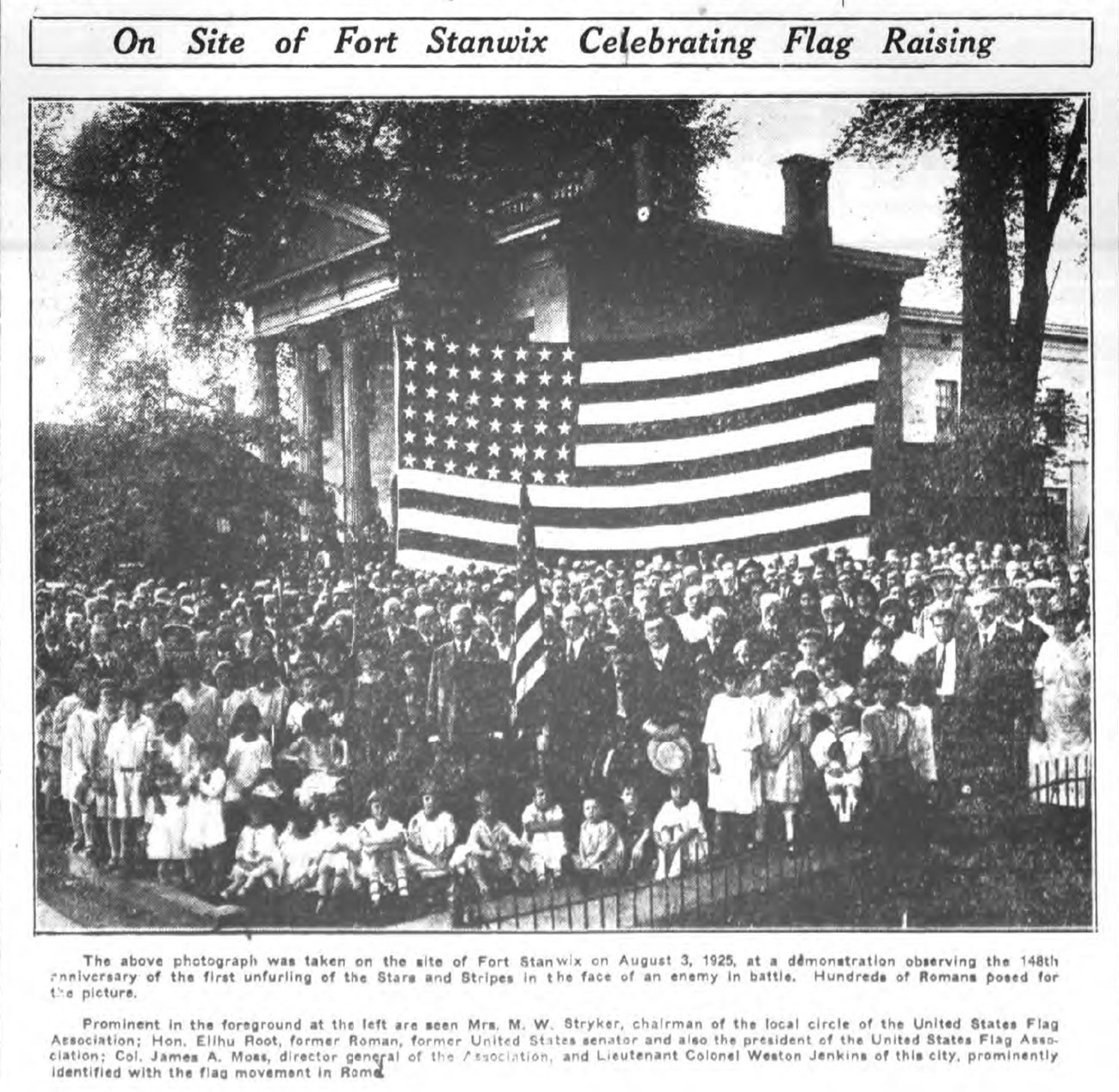 An old black and white photo of a group of children standing underneath a large American flag, in front of a large building with columns.