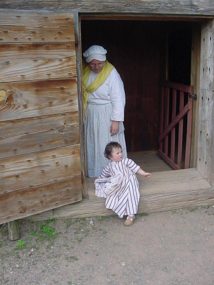A woman in 18th Century-style clothing and her baby stand in a doorway.