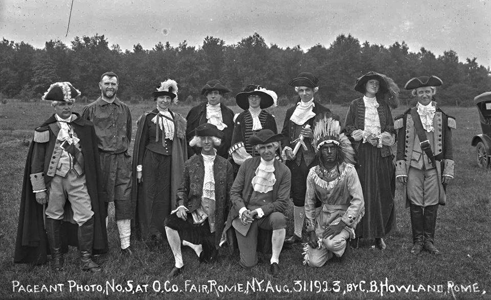 Black & white photo. Approx 15 people stand and kneel in loose rows. They are dressed in costumes to represent the American colonial period.