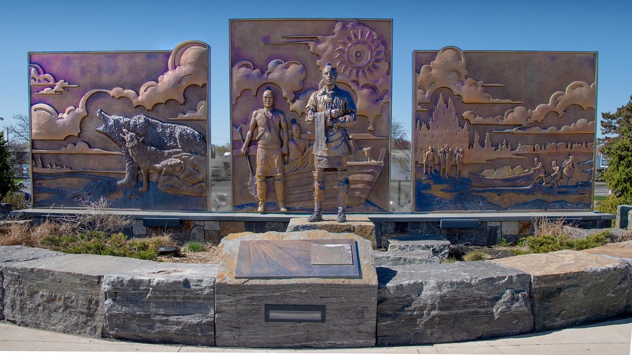 3 shiny brass panels with animals and natives in relief. In front, an Oneida man stands with feathers adorning his head and a wampum belt in hand.