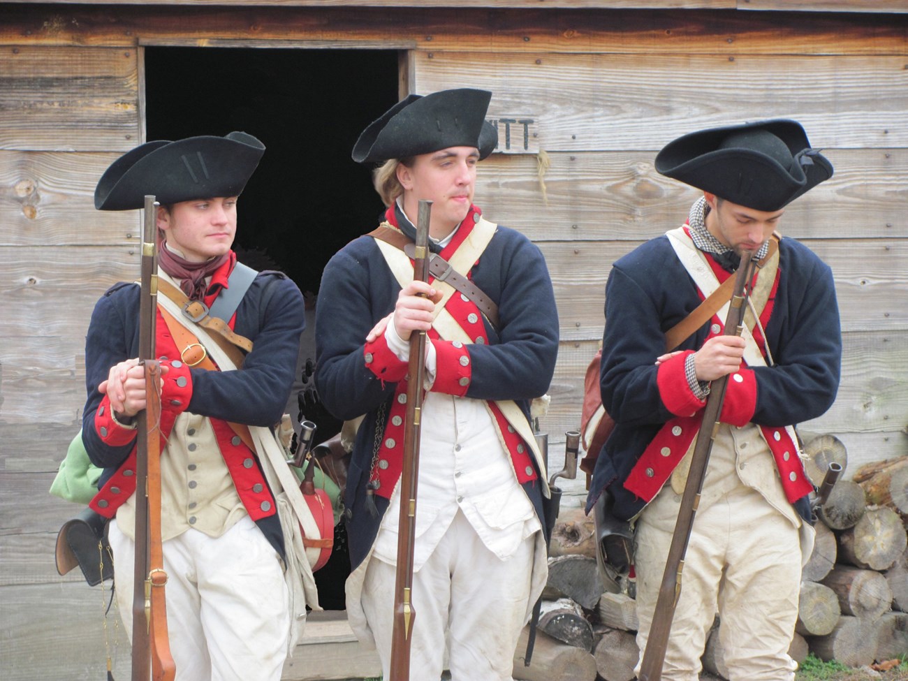 Three young Continental Soldiers stand in a row, at ease, their muskets rested in front of them