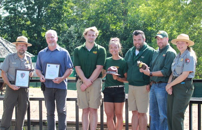 Seven people stand shoulder to shoulder in various uniforms. They hold gifts for the baby bison to show the camera.