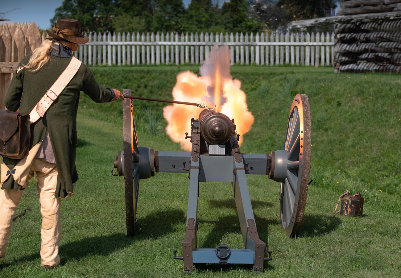 Photo of a man in a Continental uniform. From behind, he stands by a cannon with a wave of fire near the muzzle.