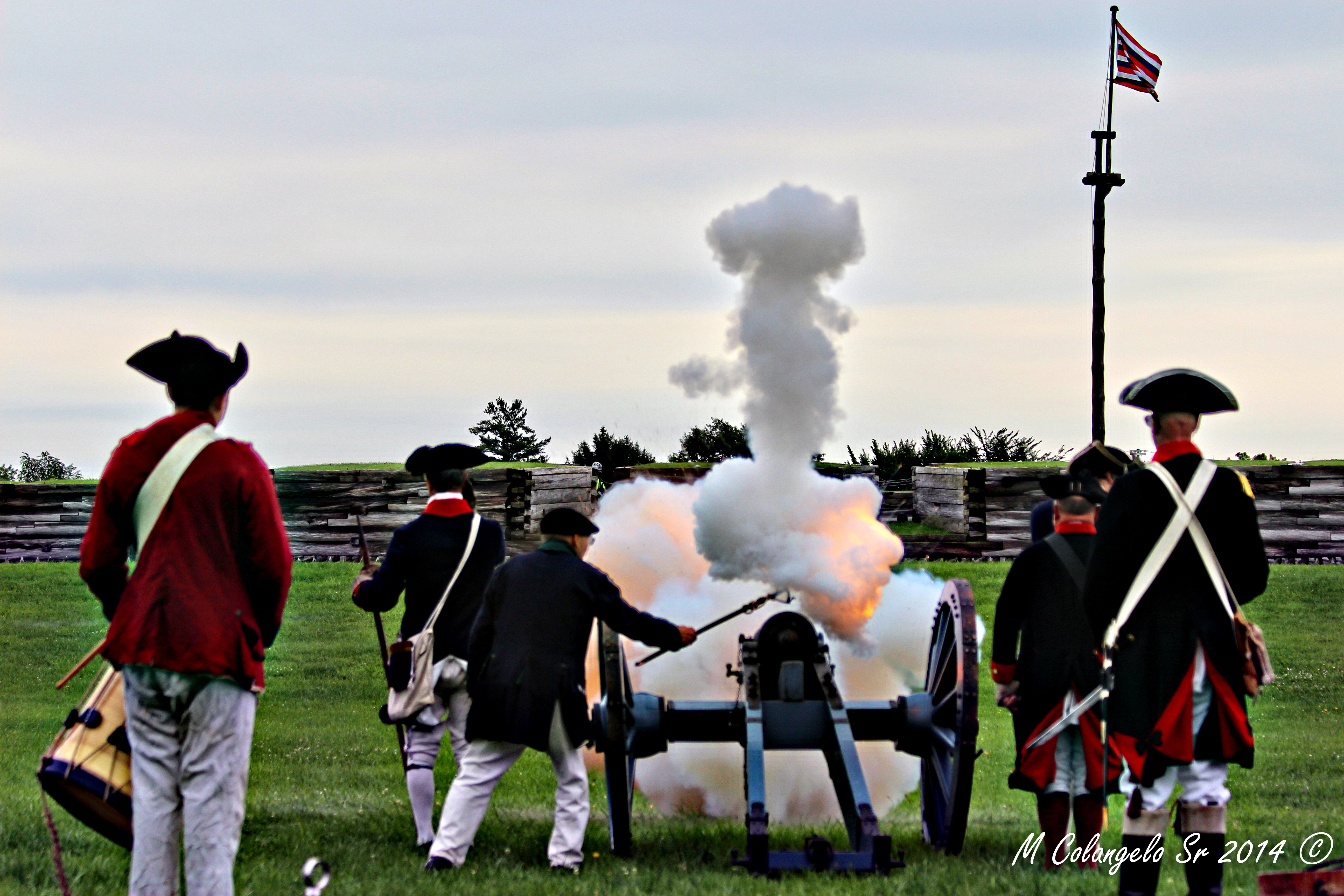 Looking at the fort in the distance, a crew of about 6 cannoneers aims at the fort. A cloud of smoke explodes around them.