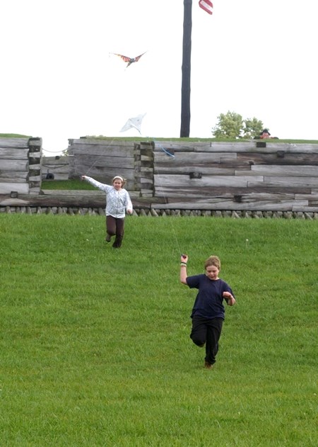 Two children run down a green grass slope outside the fort wall pulling kites behind them.