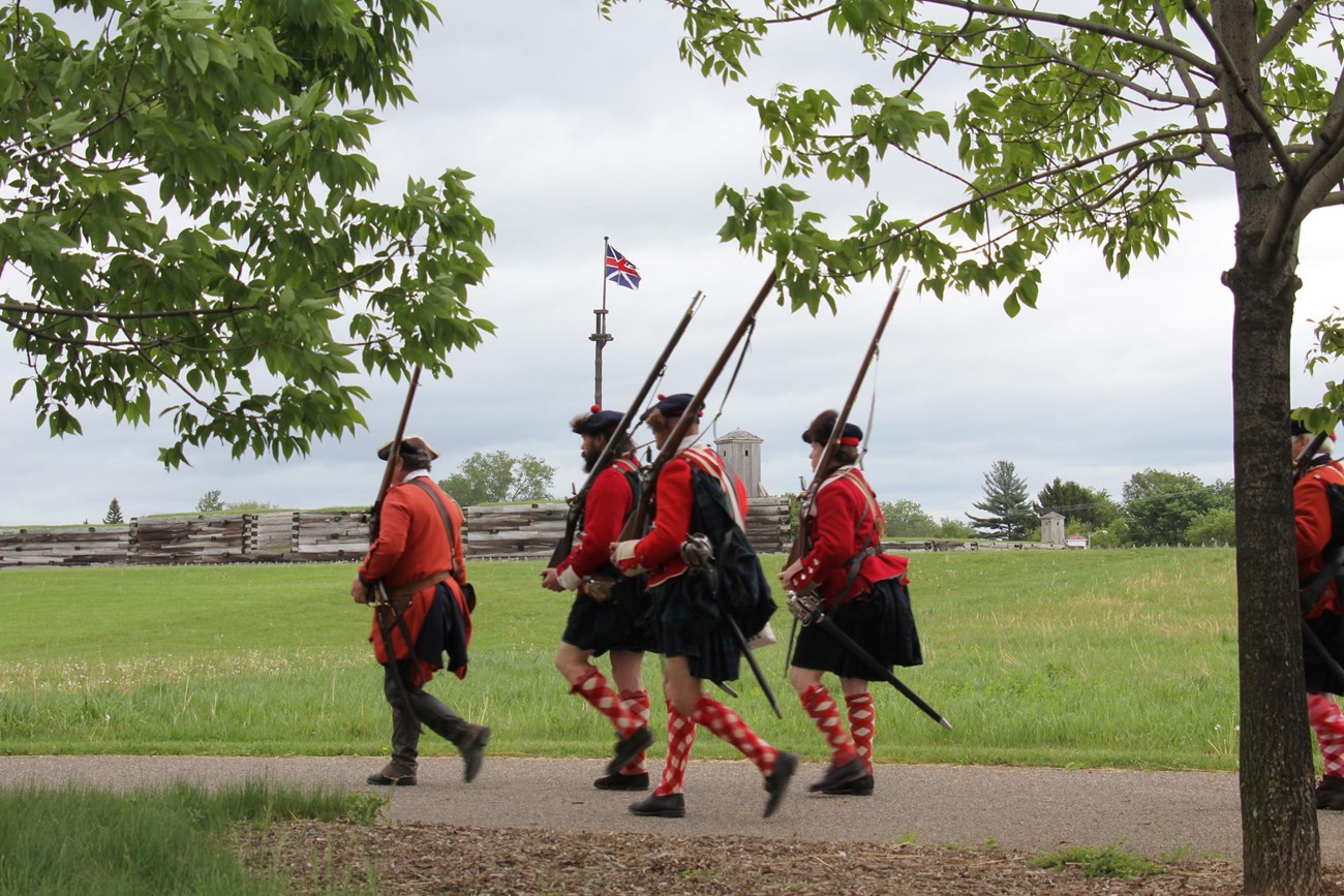 Men in varying red uniforms march outside the fort wall past the British flag.