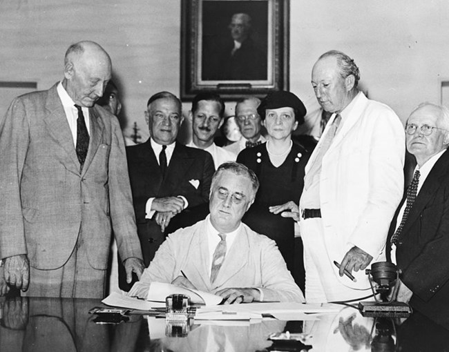 Signing_Of_The_Social_Security_Act Black and white photo of 3 men seated or standing behind a desk. They smile and hold papers.