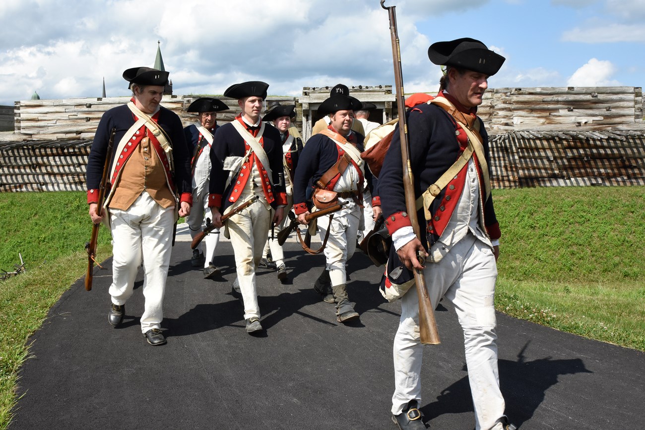 A line of Continental soldiers march in formation from the fort gate.