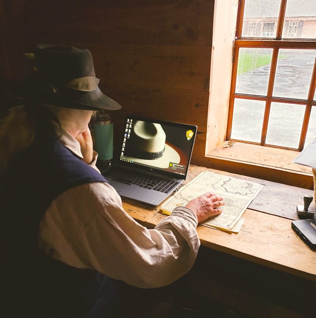 A man in 18th C clothing sits at a wooden table inside a fort room. Over his shoulder you see him looking at a modern laptop computer.