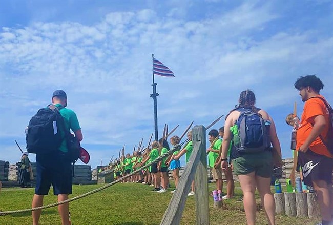 A row of little children standing in a line diagonal of you. All hold wooden muskets on their shoulders. A flag flies over their heads.