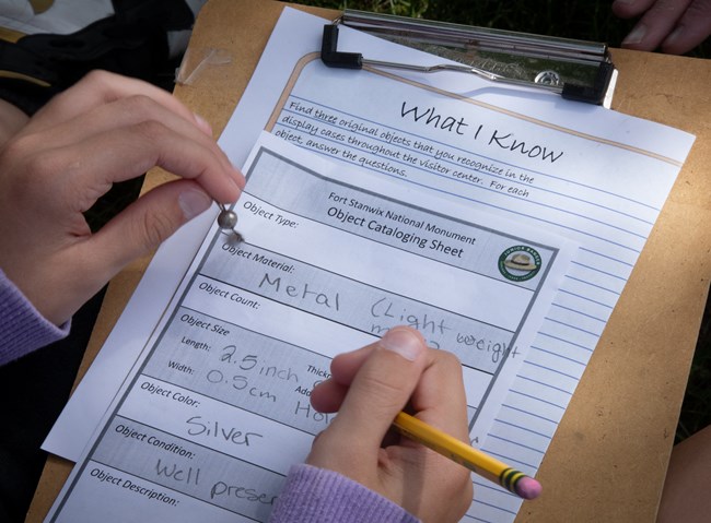 A close-up of a set of hands howling a clipboard with a worksheet on it. A pair of fingers grasp a small metal object.