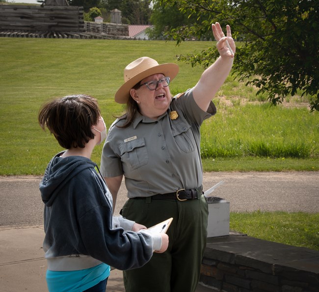 A park ranger, arm outstretched, holds an item up while a child looks on.