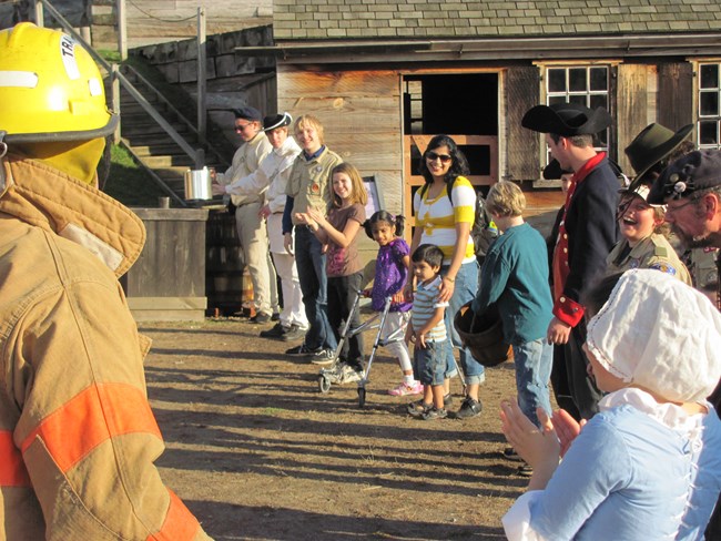 Children stand in a line passing several metal buckets back and forth.