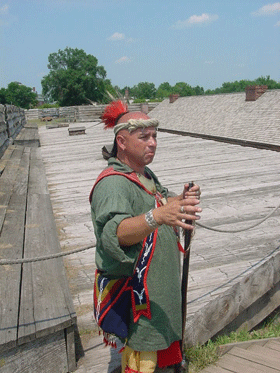 an indian warrior in a green shirt holds a rifle and gestures towards a field