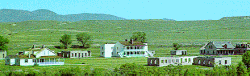 A baby blue sky over a green field, white buildings sit on the meadow