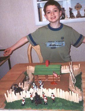 a boy with a green shirt opens his arms and smiles wide. he is showing the camera a model fort that was made by hand!