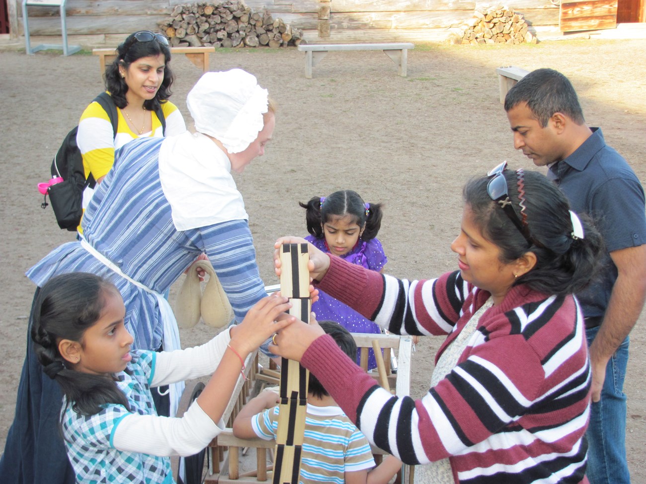 A little girl and a woman stand holding a folding wooden toy between them. The rest of their family plays behind them.