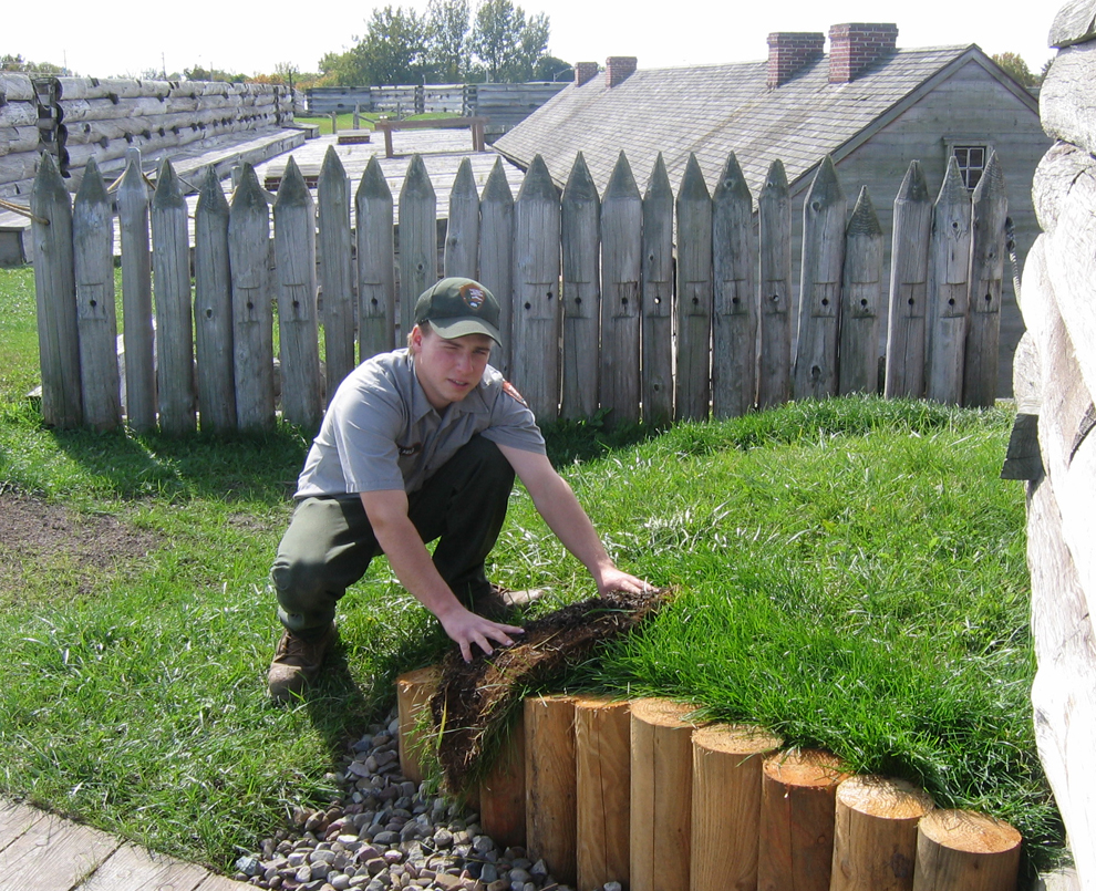 a man in a grey shirt crouches down and places green sod near a wooden surface