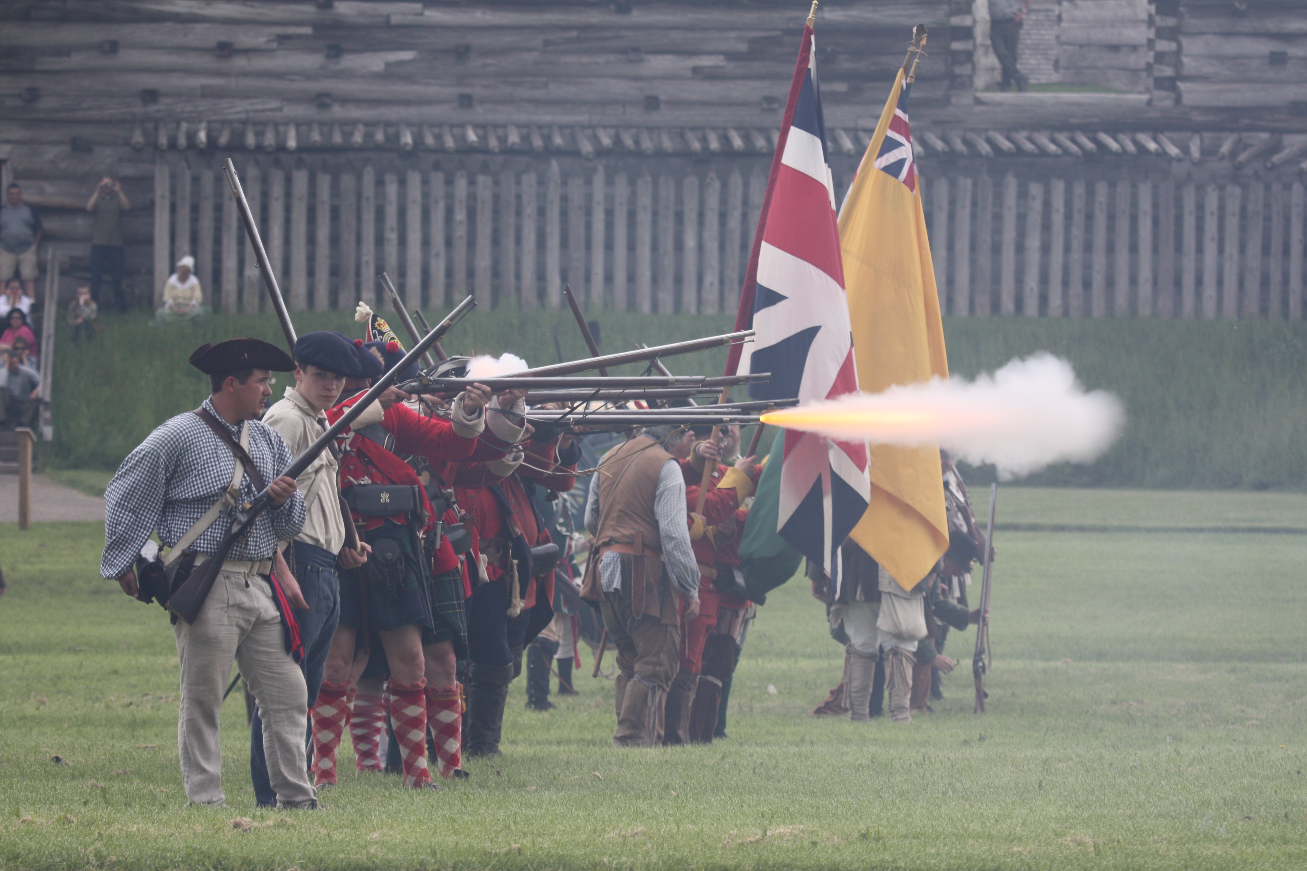 Soliders march in a line with a multi-colored flags. Smoke billows in from of them, pouring from muskets.