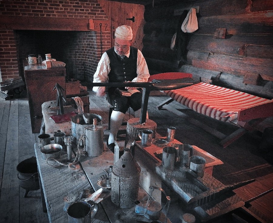 A man in 18th century clothing sits with an anvil in front of him. It is a large black piece of metal. On that he hammers a shiny piece of silver gently.