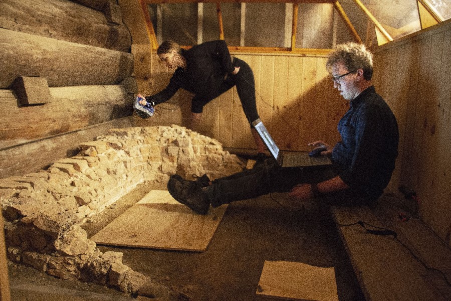 A man sits in a small wooden box with a laptop on his lap. He is trying to squeeze himself into a corner. In front of him, a woman leans over a square of crumbling bricks. She points a bulky metal gun-like object at them and carefully steps around the box