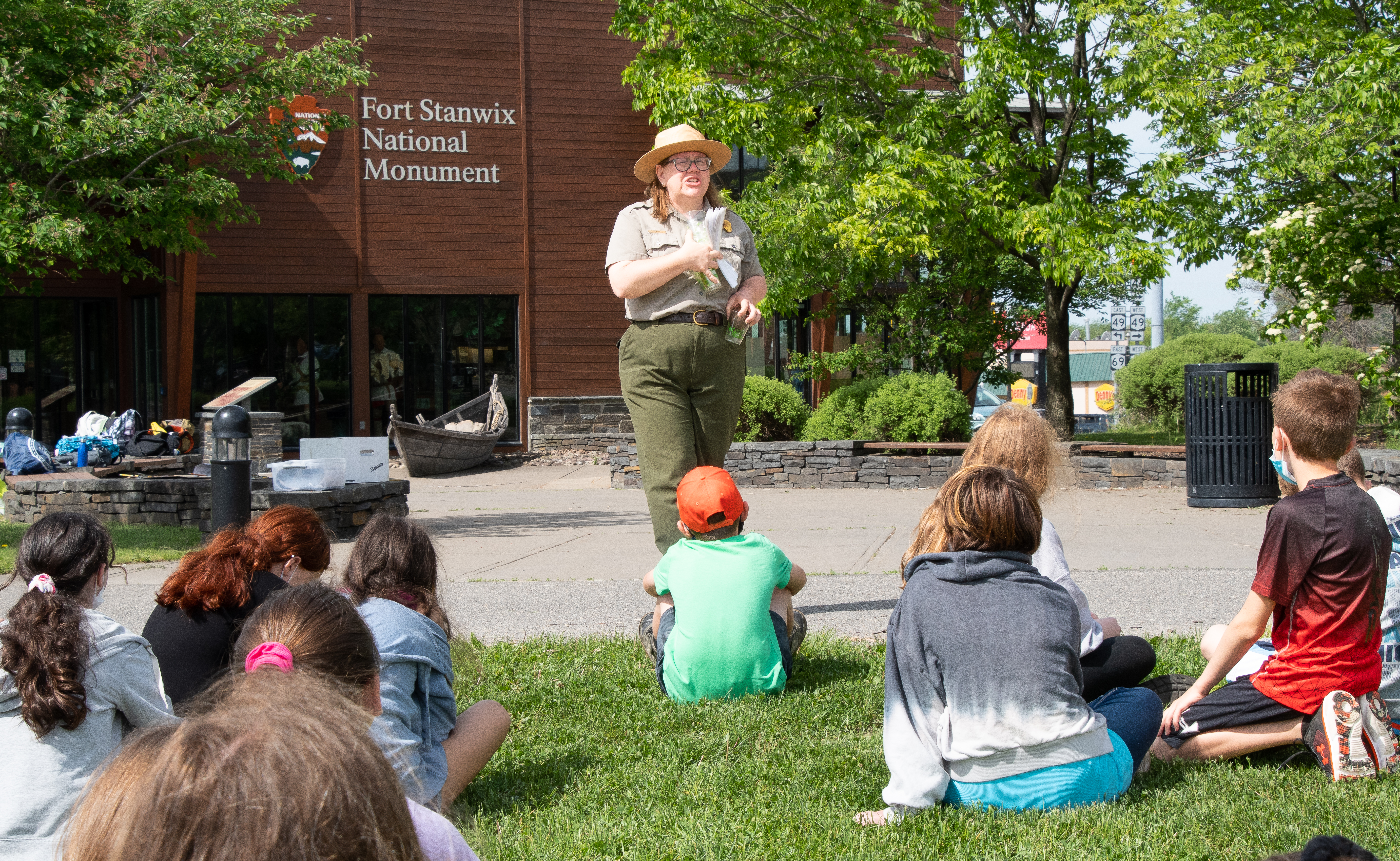 DSC_9407 A park ranger stands in front of a group of children seated on the ground in a very verdant area. You are looking at the ranger over their heads.
