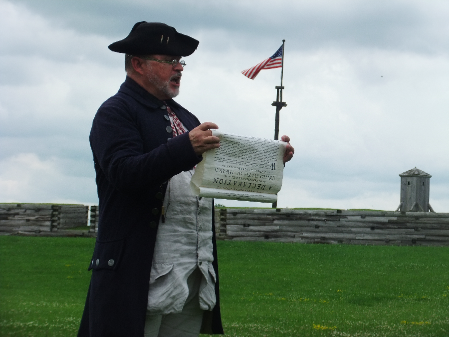 FOST04July2013 011.900pix.JPG A man in a blue 18th century style jacket with a black cocked hat. He holds a paper scroll with elaborate lettering on it. He is reading aloud from the scroll.
