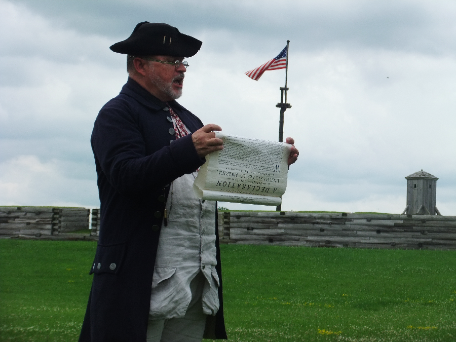 A man in a blue 18th century style jacket with a black cocked hat. He holds a paper scroll with elaborate lettering on it. He is reading aloud from the scroll.