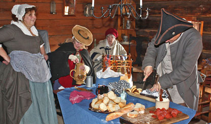 several busy ladies gather around a colorful table preparing a feast. a man with his face down cuts loafs of bread