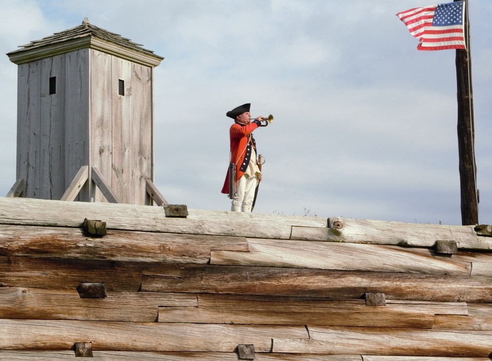 A man in a bright red jacket stands on a wooden wall playing a bugle.