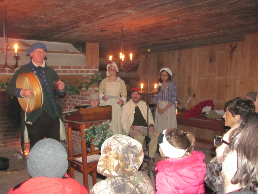 A group of people in winter coats huddle in front of 18th century carolers near a fire.