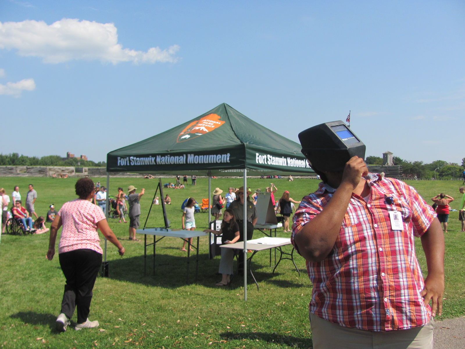 A man stands staring towards the sunny, clear sky, a welding mask over his head. Behind him a park ranger speaks to a group of people under a tent.