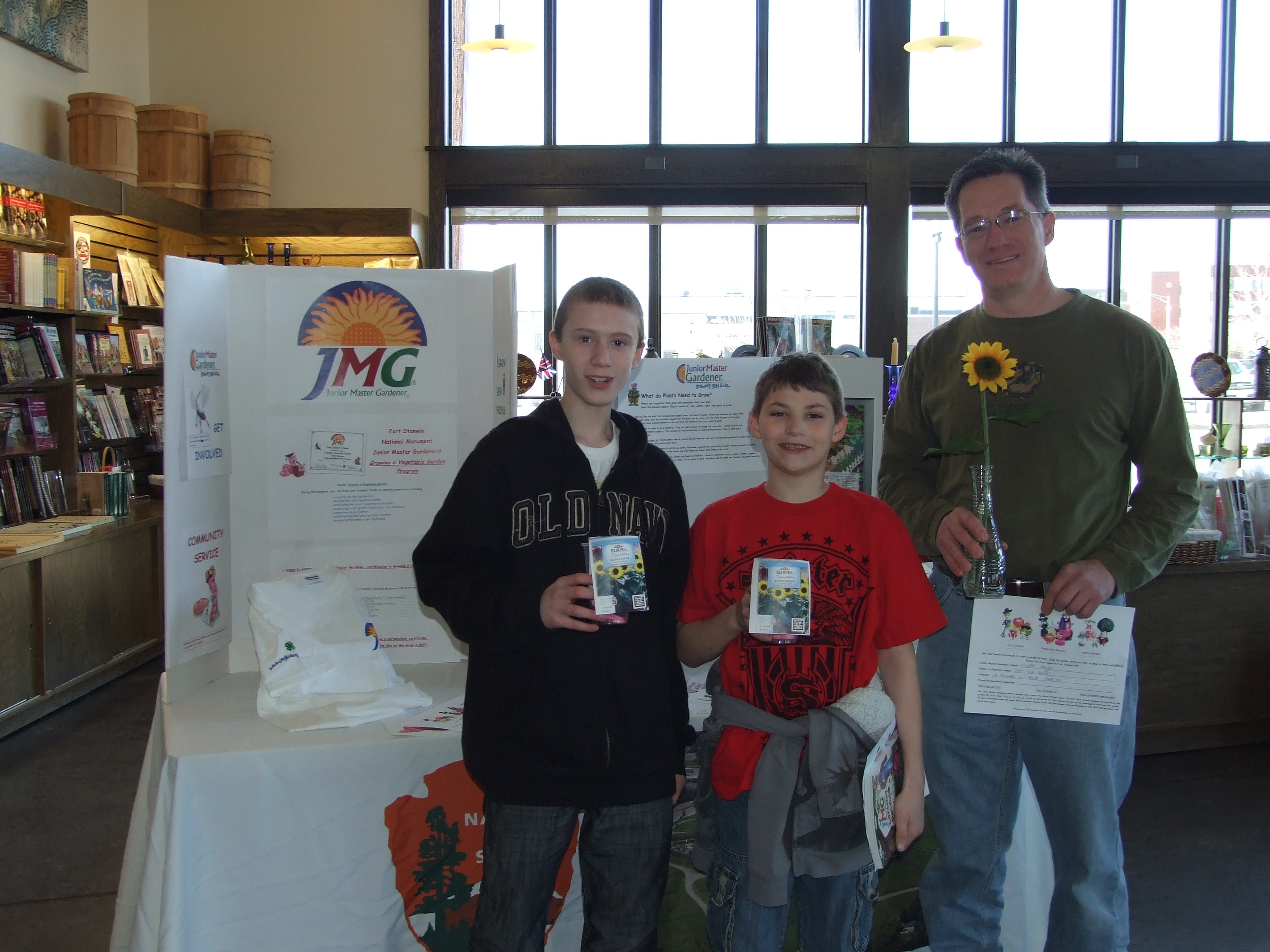 two boys and an adult stand holding plants in front of a colorful poster