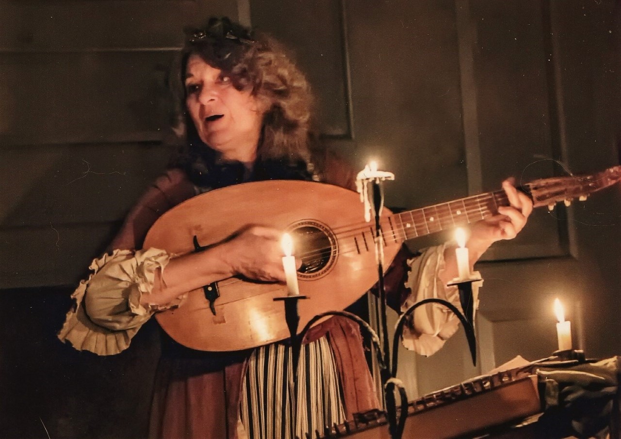 A woman in 18th Century-style clothing stands, illuminated by candle-light and playing a guitar.