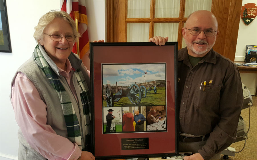 A man and a woman stand smiling.They hold a framed collage between them. The man's is featured in the collage photos.