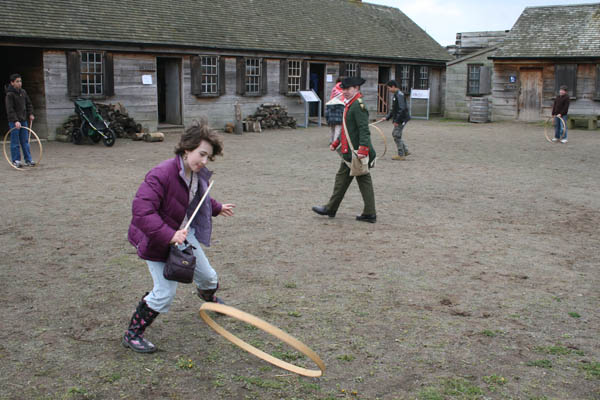 a child in a purple jacket runs past a soldier while chasing a wooden hoop.