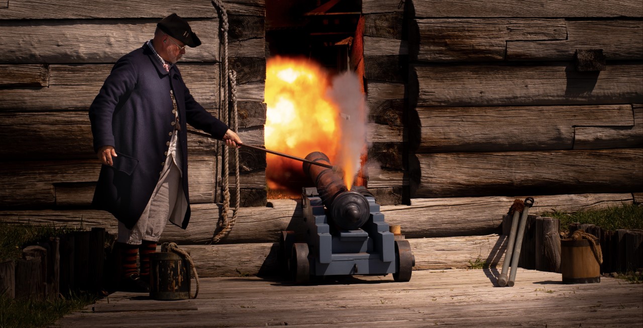 thumbnail_D85_3936 A man stands over a cannon that is facing away from you. He is surrounded by fire and smoke.