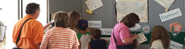 adults and children in bright clothing stand near a panel with push pins and charts