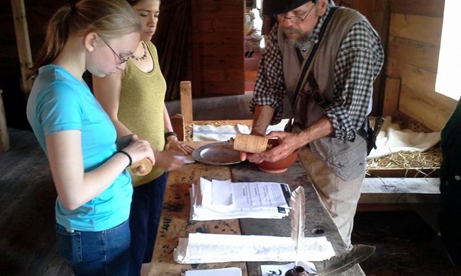 A girl and a soldier stand leaning over a table covered in papers. The man holds a wooden cylinder for her to see.