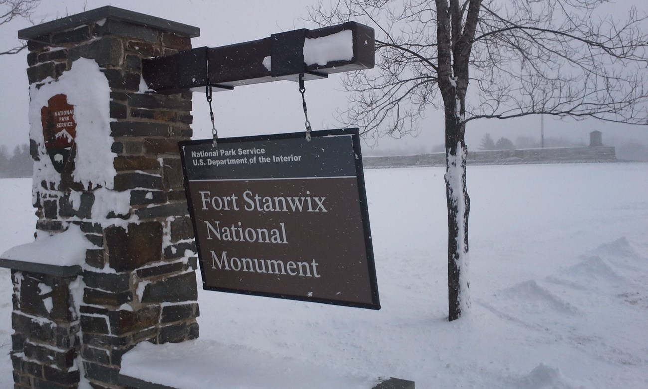 Snow covers the park sign. The wind is so strong the sign leans in the breeze.