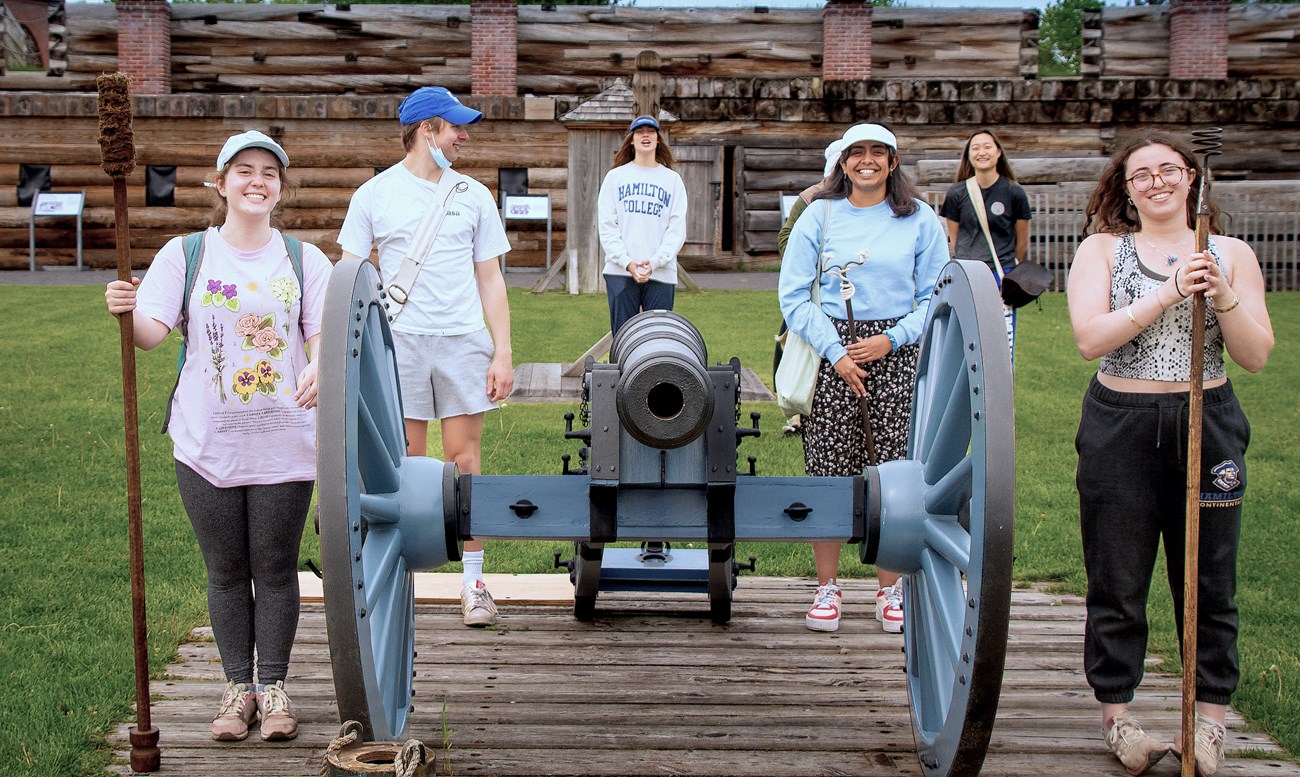Six women stand around a cannon faced at you. They are holding various tools while they learn how it works.