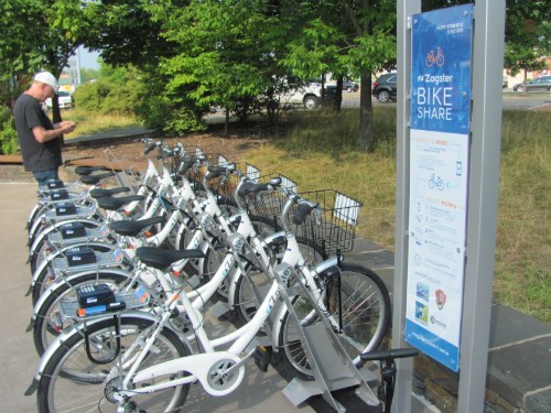 A row of white aluminum bicycles attached to a metal rack. A man stands looking over them. A row of white aluminum bicycles attached to a metal rack. A man stands looking over them.
