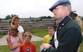 A tall soldier wears a green woolen jacket, he is speaking to children in multicolored tee-shirts on a corner of the fort. A tall soldier wears a green woolen jacket, he is speaking to children in multicolored tee-shirts on a corner of the fort.
