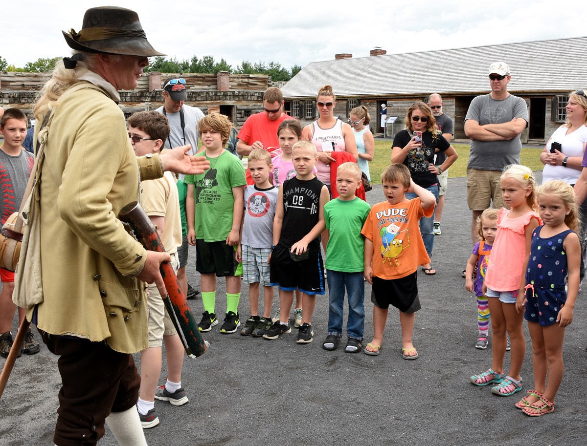 A tall Continental Soldier stands speaking in front of a crowd of children with their parents behind them.