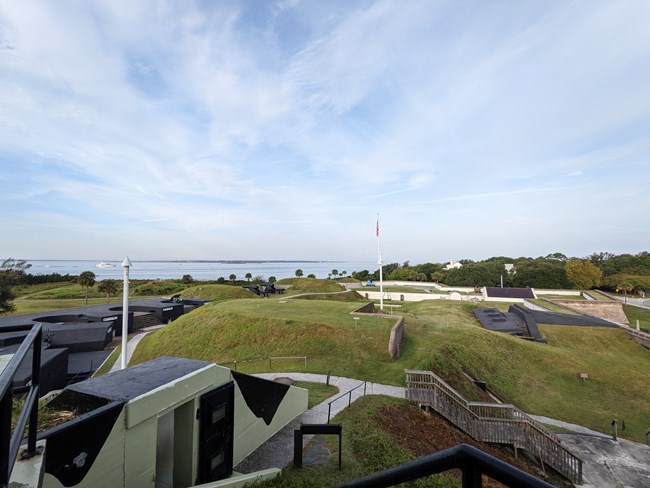 Interior of Fort Moultrie showing an American Flag on a flagpole, cannons in the distance and lots of grassy, hilly areas.